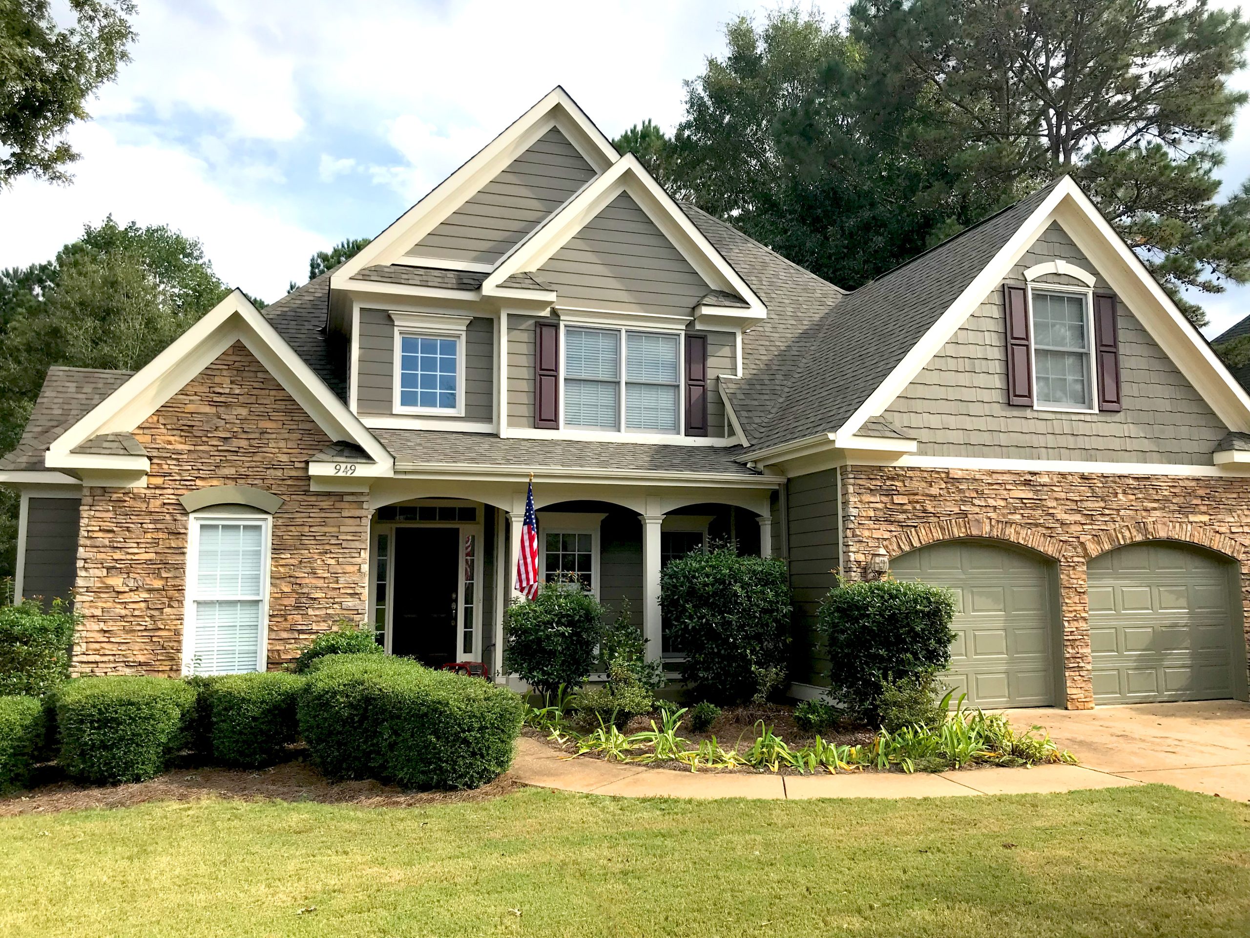 Front exterior of a two-story house with gray siding, stone accents, a dark shingle roof and an attached two-car garage, featuring a covered front porch with columns, landscaped shrubs and an American flag displayed by the entry.