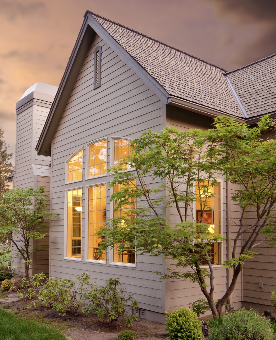 Side exterior of a light gray, two-story house with a steep gabled roof, large multi-pane windows glowing with warm interior light and landscaped shrubs and small trees at dusk.