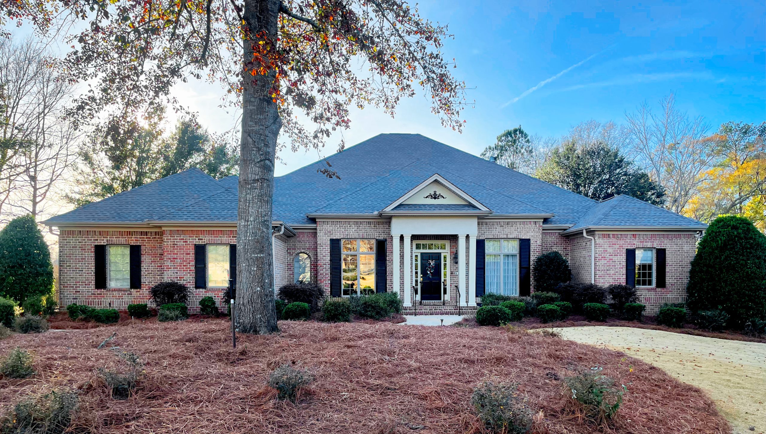 Front exterior of a single-story brick house with a dark gray hip roof, black shutters and a centered covered entry with white columns, surrounded by trimmed shrubs, pine straw landscaping and a large tree in the front yard under a clear blue sky.